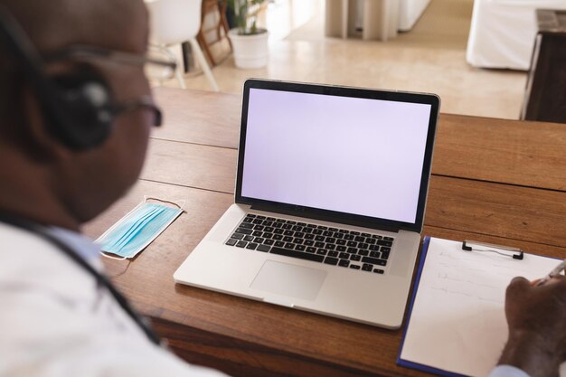 Médico afro-americano sênior escrevendo na área de transferência com laptop com espaço de cópia na mesa