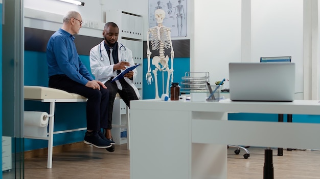 Médico afroamericano consultando a un anciano con enfermedad, dando medicamentos recetados y tratamiento para curar el diagnóstico. Médico y paciente hablando sobre asistencia sanitaria en el gabinete.