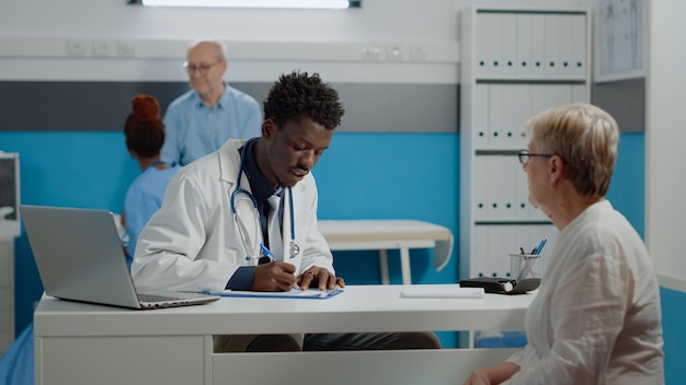 Médico afroamericano escribiendo chequeo en el cuaderno mientras consulta a la mujer mayor en el escritorio. Médico negro comprobando la atención médica del paciente mientras la enfermera y el anciano hablando en segundo plano.