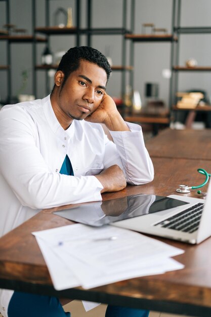 Un médico afroamericano estresado mirando una laptop leyendo malas noticias en línea preocupado por un error en el lugar de trabajo. Médico infeliz en tensión sintiéndose cansado resolviendo problemas informáticos en el trabajo.