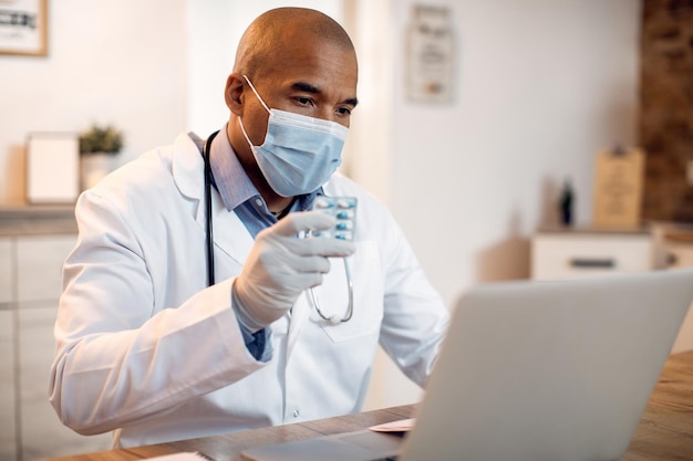 Médico afroamericano con mascarilla que aconseja a un paciente sobre el medicamento a través de una videollamada desde su oficina