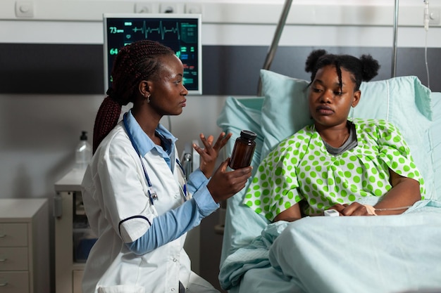 Médico afroamericano médico de pie junto al paciente enfermo sosteniendo una botella de analgésico explicando el tratamiento médico durante el examen médico en la sala del hospital. Servicios de medicina