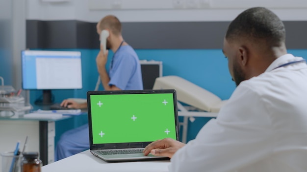 Médico afroamericano mirando simulacro de computadora portátil con pantalla verde chroma key con pantalla aislada. Hombre terapeuta analizando diagnóstico de enfermedad trabajando en informe médico en la oficina del hospital