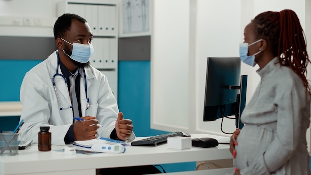 Médico afroamericano y paciente con embarazo hablando sobre el parto, haciendo consultas en el escritorio de la oficina. Médico y futura madre conversando durante la pandemia de covid 19.