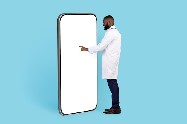 Médico afroamericano parado cerca de un gran teléfono inteligente en blanco y tocando la pantalla blanca