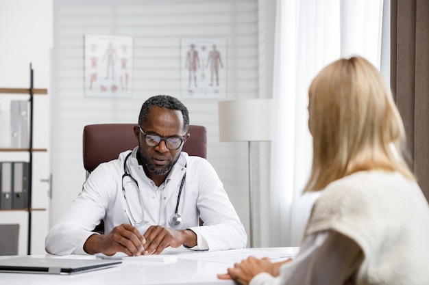 Médico afroamericano recibe un paciente escucha quejas hace diagnósticos escribe datos docto
