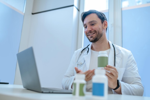 Médico amigável sentado à mesa segurando um frasco plástico de suplemento nutricional diante da tela do laptop durante a consulta on-line