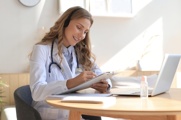 Médico amigável trabalhando online com um laptop sentado em uma mesa em uma consulta.