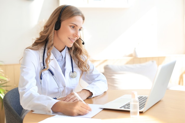 Médico amigável trabalhando online com um laptop sentado em uma mesa em uma consulta.