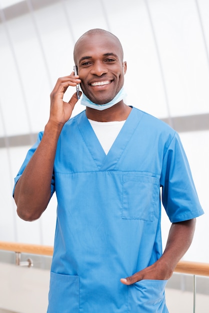 Médico ao telefone. Jovem médico africano alegre de uniforme azul falando ao celular e olhando para a câmera