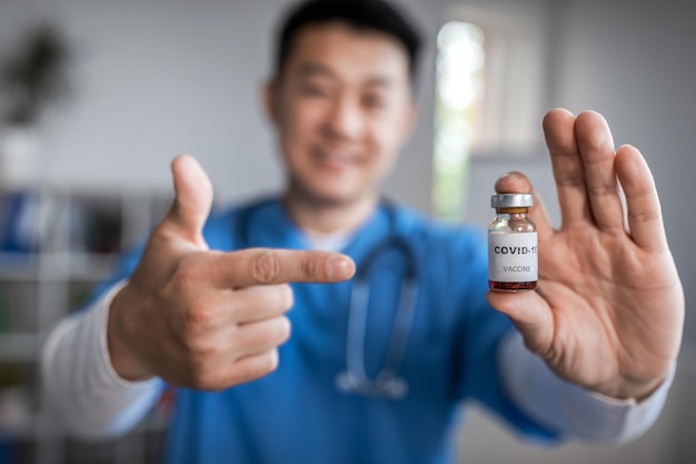 El médico asiático maduro alegre señala con el dedo la vacuna en el interior de la oficina de la clínica Lucha con la enfermedad