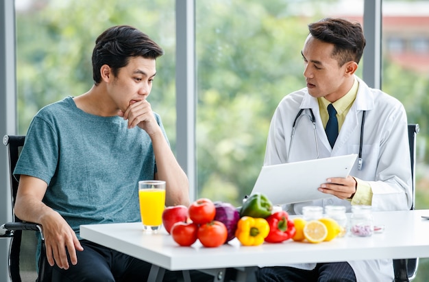 Médico asiático positivo sonriendo y demostrando la mesa al paciente masculino mientras está sentado en el escritorio con comida saludable en la clínica