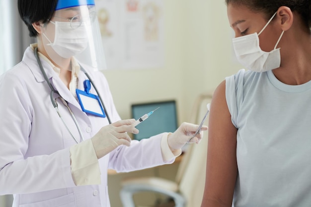 Médico asiático con ropa protectora inyectando a una adolescente en el brazo durante un examen médico en el hospital