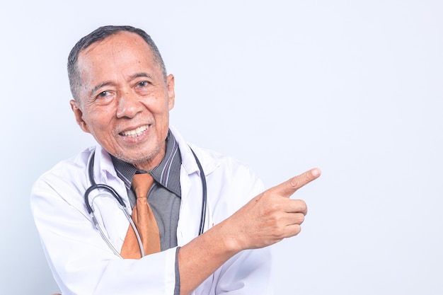 Médico asiático sênior com estetoscópio apontando para o espaço vazio enquanto olha para a câmera sorrindo