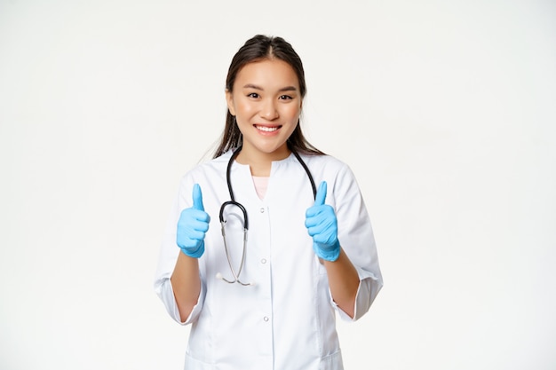 Médico asiático sorridente, mulher em uniforme médico e luvas de borracha mostra os polegares, recomendando smth ao paciente, em pé sobre um fundo branco.