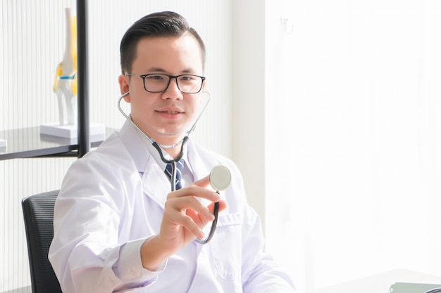 Un médico asiático, tailandés, lleva gafas y uniforme. El médico está sentado, sosteniendo y sosteniendo el estetoscopio en la sala de examen.