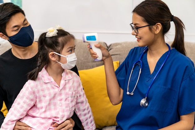 Médico asiático visita a casa y toma la temperatura a una niña en la sala de estar