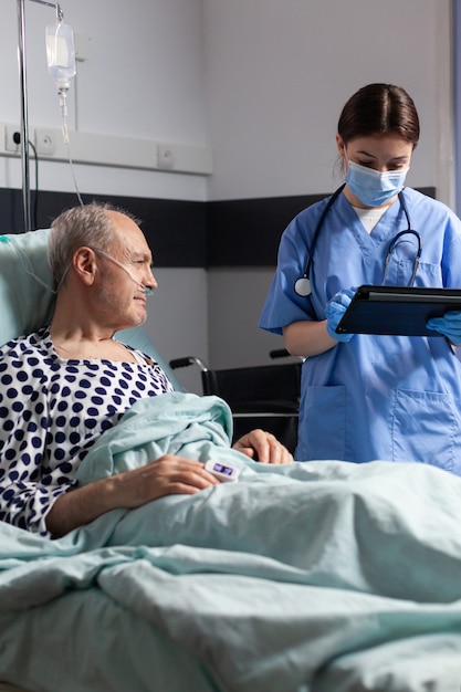 Médico assistente com máscara de proteção verificando tratamento de homem sênior lendo notas no tablet pc