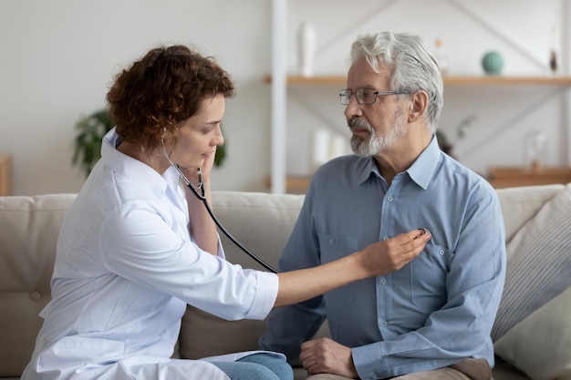 Foto médico assistente feminino segurando estetoscópio ouvindo paciente idoso durante visita domiciliar médico verificando batimentos cardíacos examinando idoso aposentado em casa conceito de cardiologia de doenças cardíacas de idosos