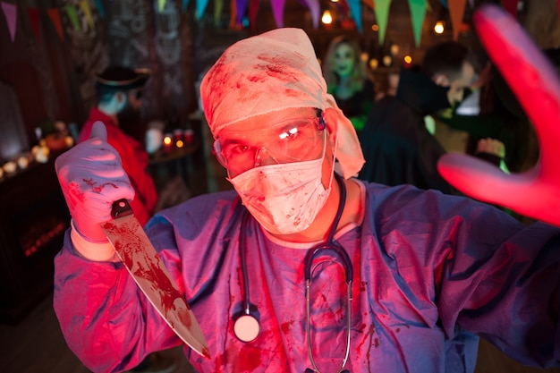 Médico assustador com seu coberto furando um kinfe com sangue e olhando para a câmera na festa de halloween. Evento de Halloween.