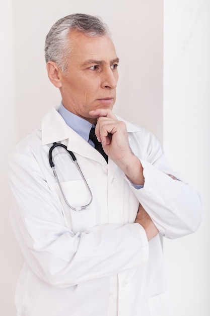 Médico atencioso. Médico sênior de cabelo grisalho de uniforme olhando para o lado e segurando o queixo