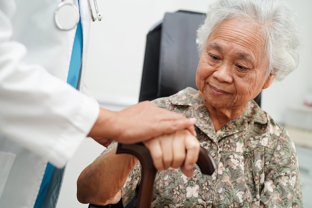 El médico ayuda a una anciana asiática con discapacidad a una paciente que sostiene un bastón en la mano arrugada en el hospital