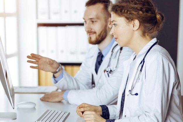 Médico de barba roja con una colega discutiendo la terapia actual de la enfermedad mientras se sienta en el lugar de trabajo en una clínica soleada. Trabajo en equipo en medicina.