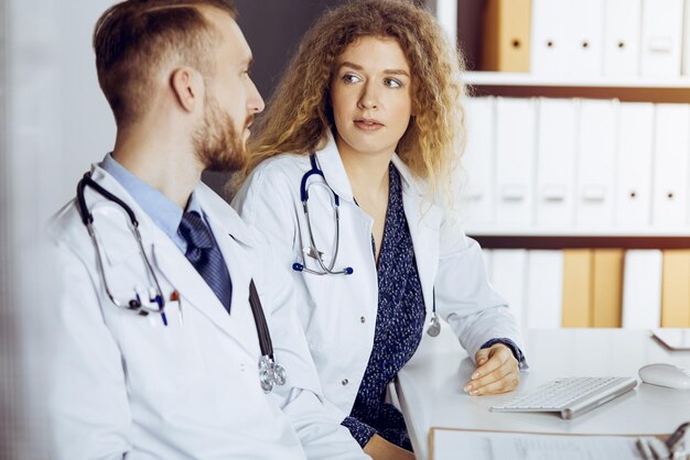 Médico barbudo con una colega discutiendo la terapia actual de la enfermedad mientras se sienta en el lugar de trabajo en una clínica soleada. trabajo en equipo en medicina