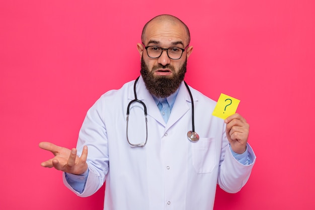 Médico barbudo com jaleco branco com estetoscópio no pescoço e óculos segurando um papel de lembrete com ponto de interrogação confuso, levantando o braço em desgosto