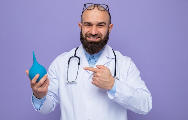 Médico barbudo com jaleco branco com estetoscópio no pescoço segurando uma pêra médica apontando com o dedo indicador para ela sorrindo alegremente em pé sobre o fundo roxo