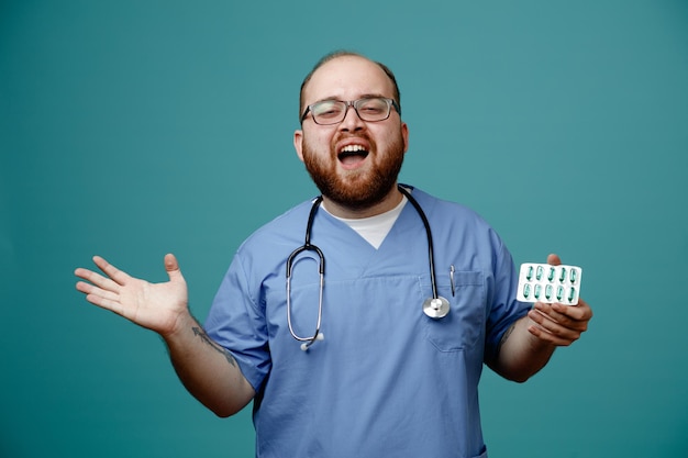 Médico barbudo de uniforme com estetoscópio no pescoço usando óculos segurando pílulas sobre fundo azul