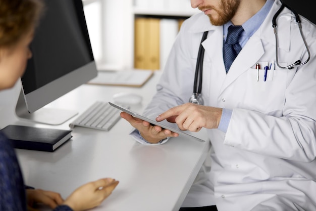 Médico barbudo desconhecido e mulher paciente discutindo o exame de saúde atual enquanto está sentado e usando o computador tablet, close-up. Conceito de medicina.