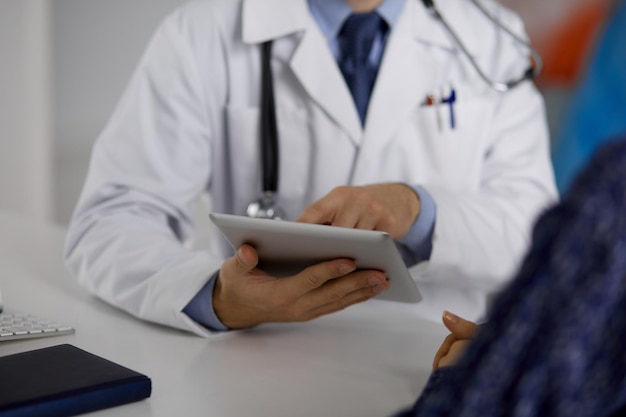 Médico barbudo desconhecido e mulher paciente discutindo o exame de saúde atual enquanto está sentado e usando o computador tablet, close-up. Conceito de medicina.