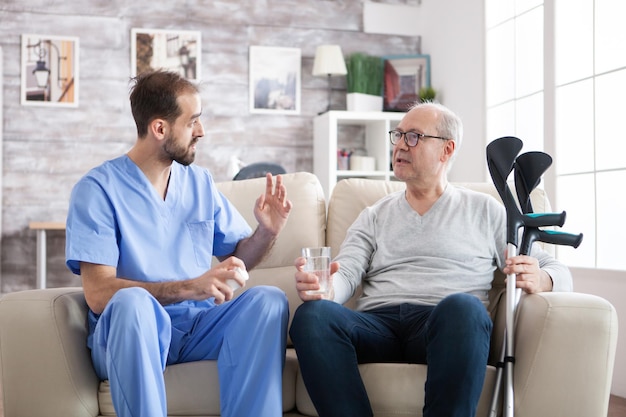 Médico barbudo en hogar de ancianos dando pastillas a pacientes mayores con muletas.