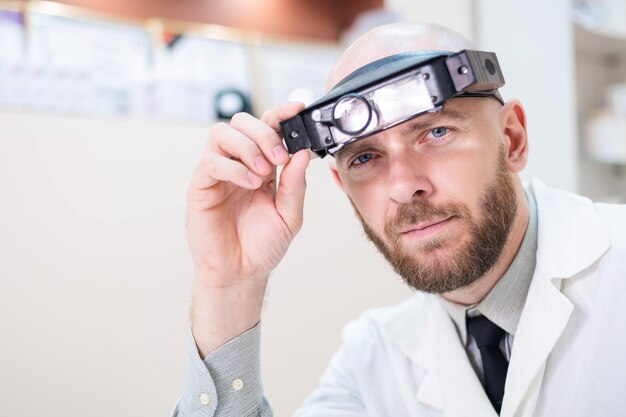 Médico barbudo masculino em um jaleco e com uma alça de cabeça de lupa w luzes sentado na mesa Equipamento de optometrista Consultório médico