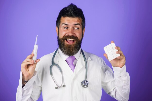 Médico barbudo con pastillas y jeringa en manos médico sonriente con estetoscopio y bata médica