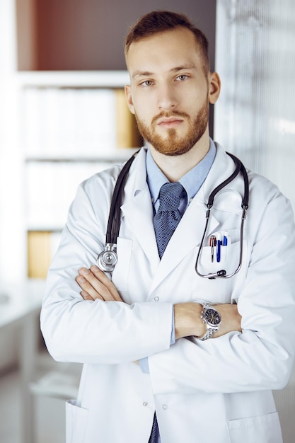 Médico barbudo rojo parado en una clínica soleada cerca de su lugar de trabajo. Retrato de médico. concepto de medicina.