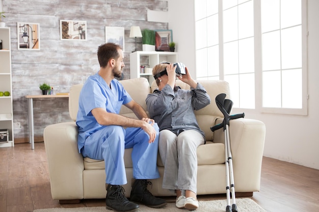 Médico barbudo sentado en un sofá en un hogar de ancianos mientras una anciana con muletas usa gafas de realidad virtual.