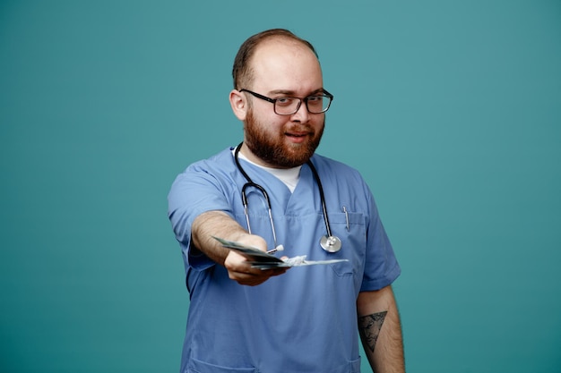 Médico barbudo en uniforme con estetoscopio alrededor del cuello con gafas con dinero en efectivo ofreciendo dinero mirando a la cámara sonriendo astutamente de pie sobre fondo azul.