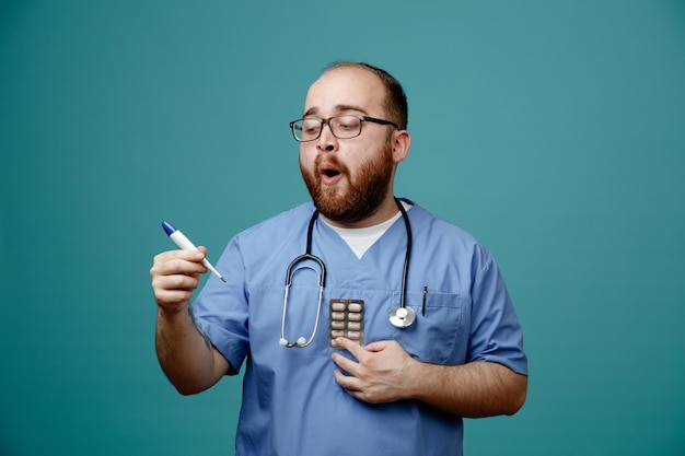 Médico barbudo en uniforme con estetoscopio alrededor del cuello con gafas con termómetro y pastillas que parecen sorprendidos y asombrados de pie sobre fondo azul.