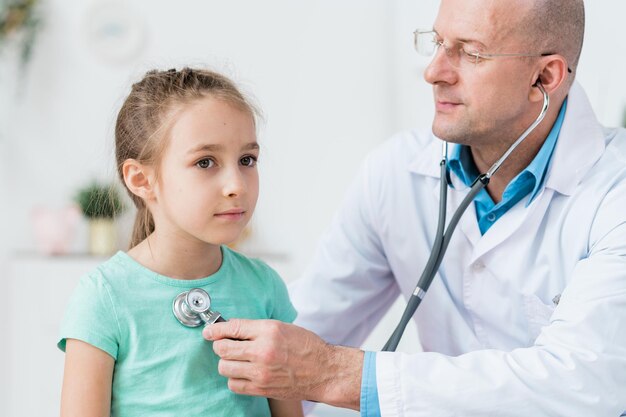 Médico con bata blanca y gafas examinando a un pequeño paciente con un estetoscopio