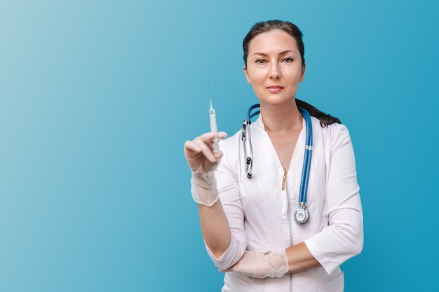 Un médico con bata blanca y guantes de goma con una jeringa mira a la cámara, fondo azul.