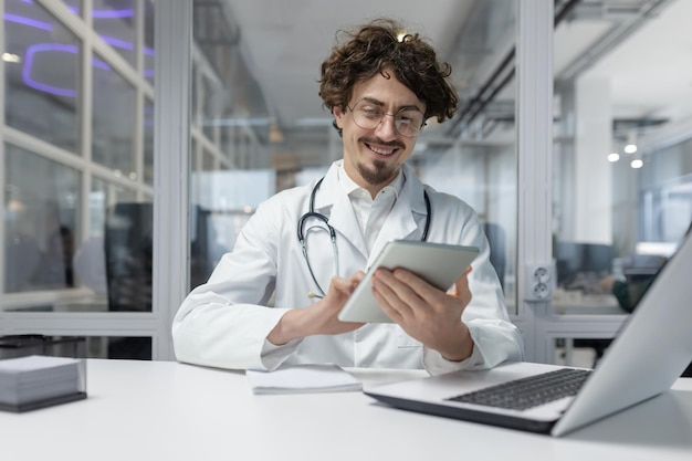 Un médico con una bata blanca de laboratorio y un estetoscopio es visto usando una tableta dentro de un consultorio médico en