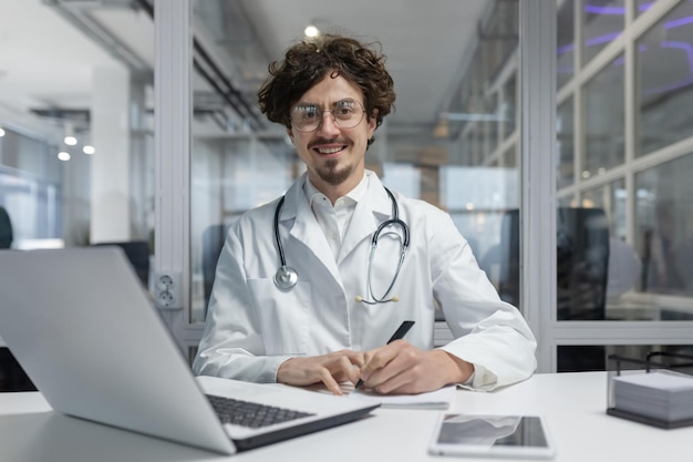 Un médico con una bata blanca de laboratorio y un estetoscopio está sentado en un escritorio usando una computadora portátil hombre sonríe feliz