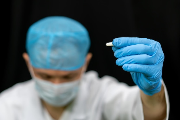 Un médico con bata blanca sostiene una pastilla en la mano sobre un fondo negro Preparaciones médicas para el tratamiento del coronavirus Un joven médico interno está considerando un nuevo medicamento