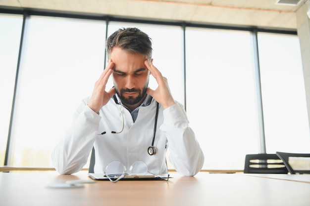 Médico bonito vestindo uniforme médico sentado em seu local de trabalho cansado segurando sua cabeça sentindo fadiga e dor de cabeça Conceito de estresse e frustração
