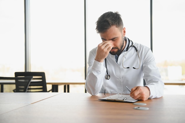 Médico bonito vestindo uniforme médico sentado em seu local de trabalho cansado segurando sua cabeça sentindo fadiga e dor de cabeça Conceito de estresse e frustração