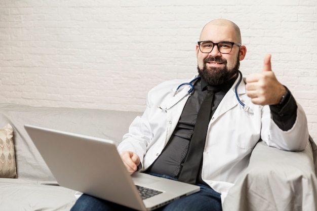 médico calvo con una bata blanca con gafas se sienta con una computadora portátil en el sofá. Consultas en línea. Cuarentena durante la pandemia de coronavirus.