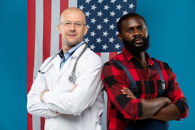 Médico calvo maduro y joven reparador africano cruzando los brazos sobre el pecho mientras te mira contra la bandera de barras y estrellas