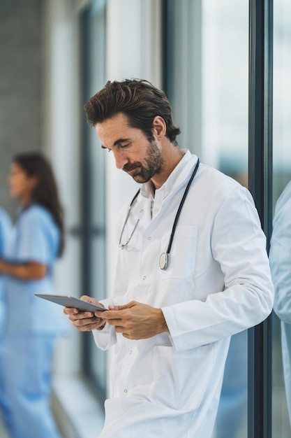 Un médico cansado que usa una tableta digital mientras está parado cerca de la ventana y tiene un descanso rápido en un pasillo del hospital.